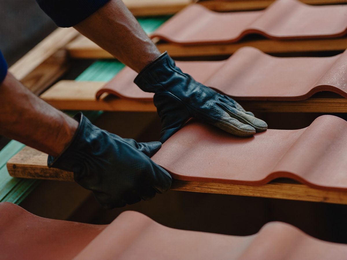 Hände in Arbeitshandschuhen legen Tonziegel auf die Dachlatten