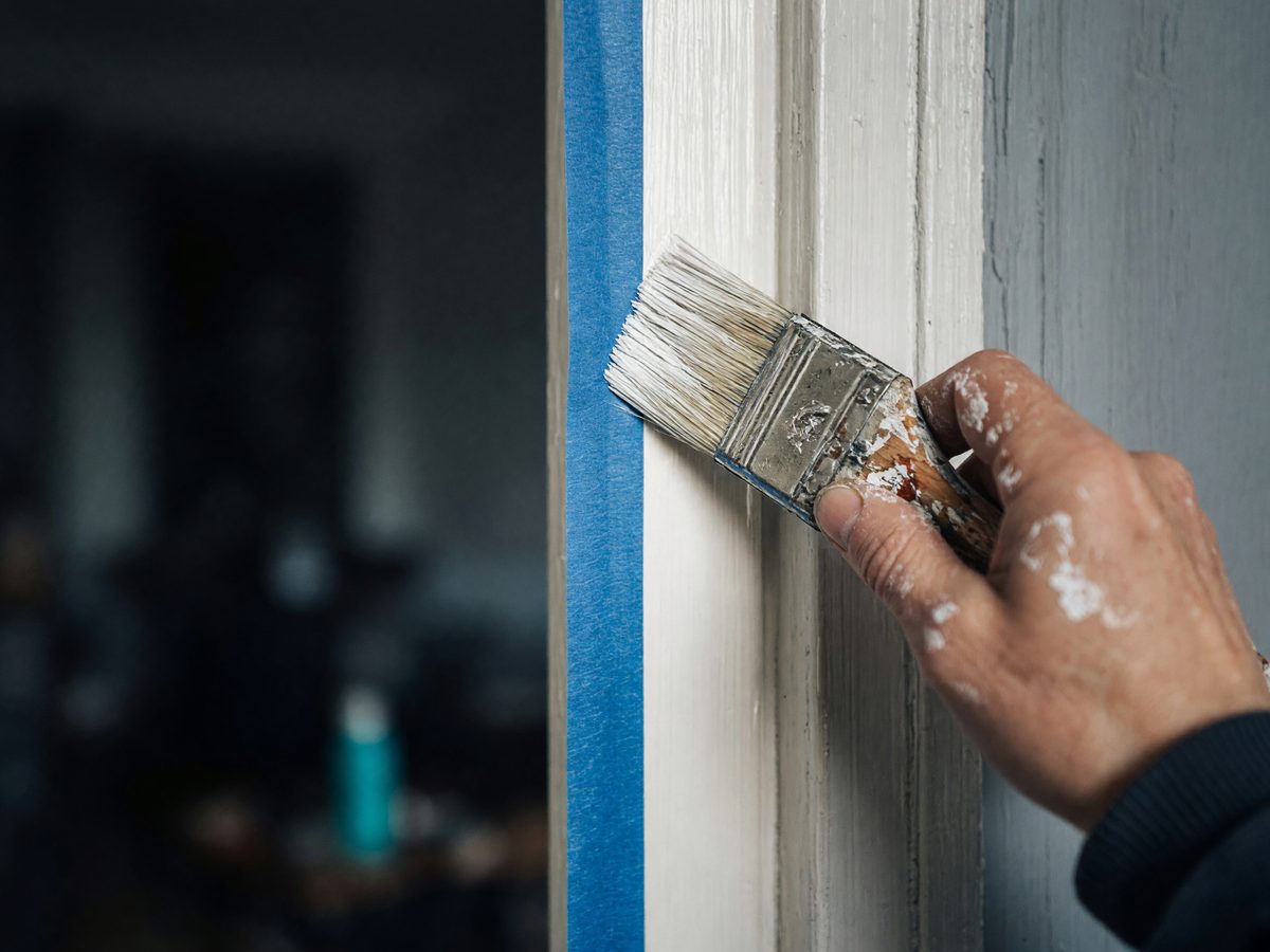 Hand mit Farbspritzern streicht einen Türrahmen entlang eines Abklebebands